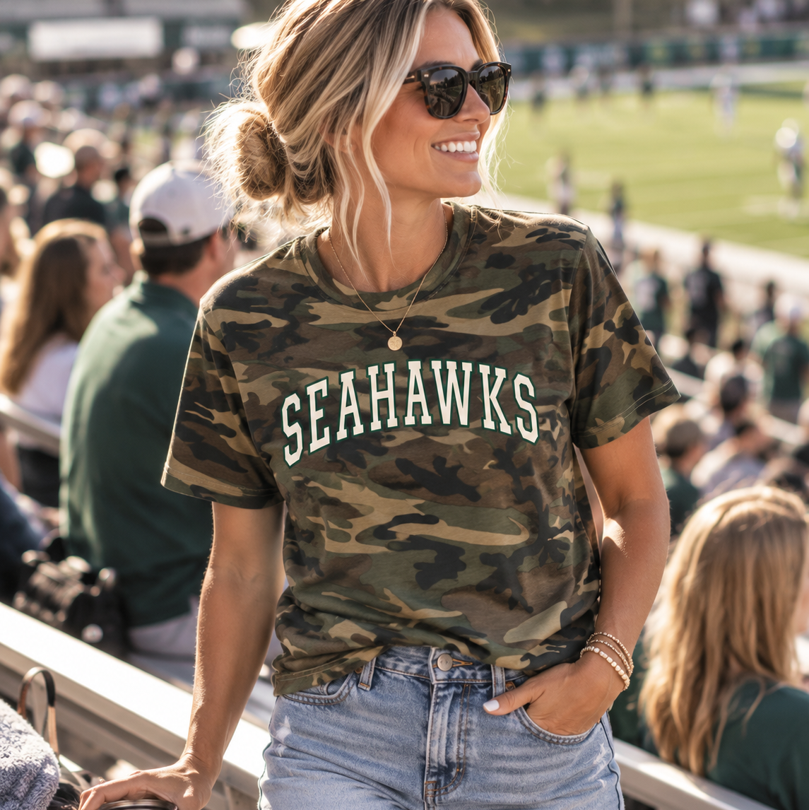 A woman wearing a teal green short-sleeve t-shirt with 'Seahawks' printed in white script across the chest, paired with denim shorts, standing in front of outdoor stadium bleachers.
