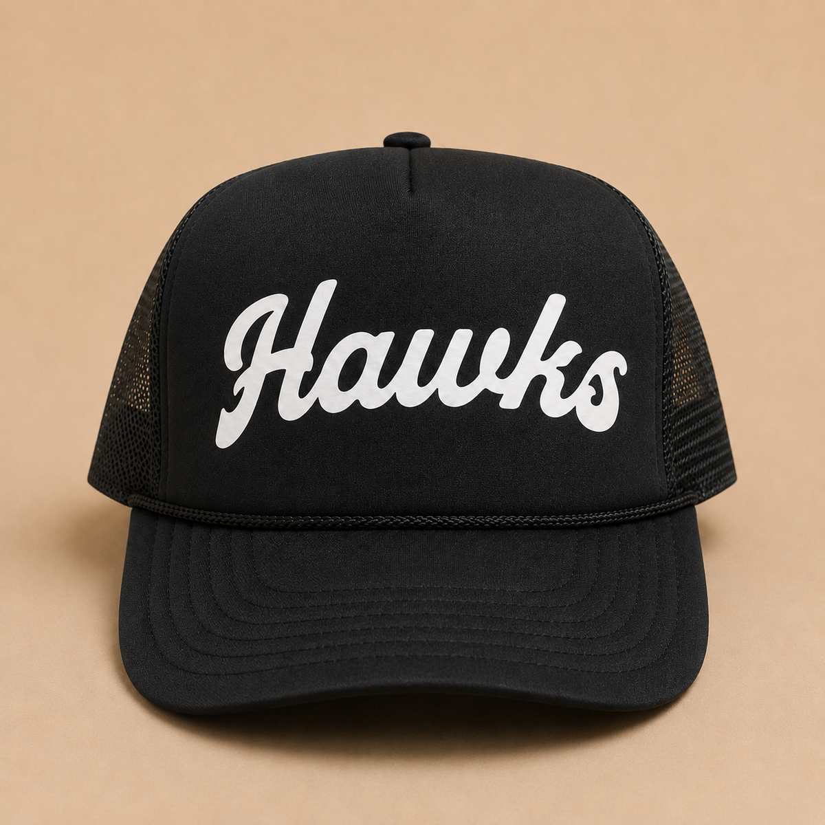 A black trucker-style cap with white cursive 'Hawks' lettering on the front, shown against a solid tan background.