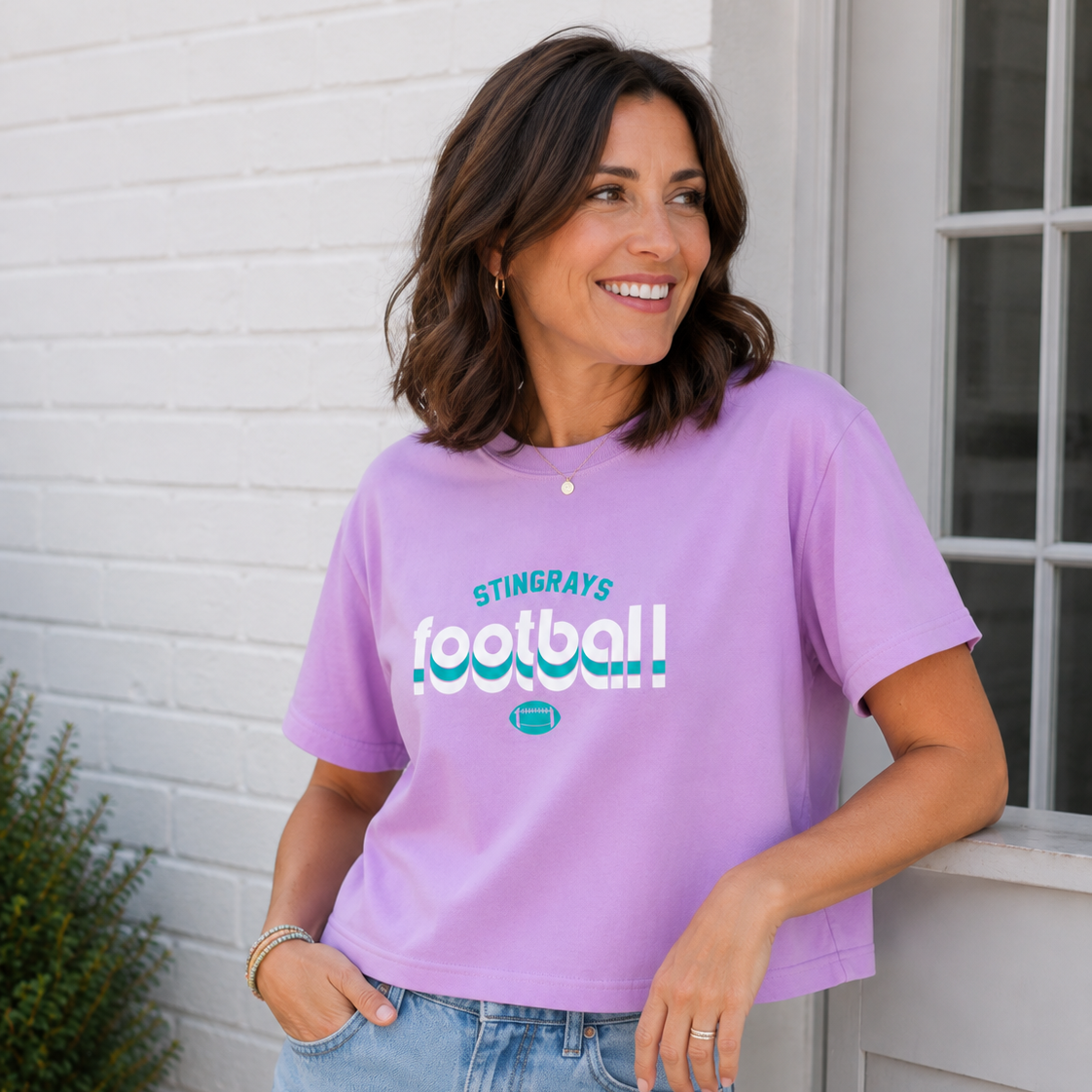 A woman wearing a light purple cropped t-shirt with 'STINGRAYS football' printed on the front in teal and white, paired with light-wash denim shorts, standing against a white brick wall.
