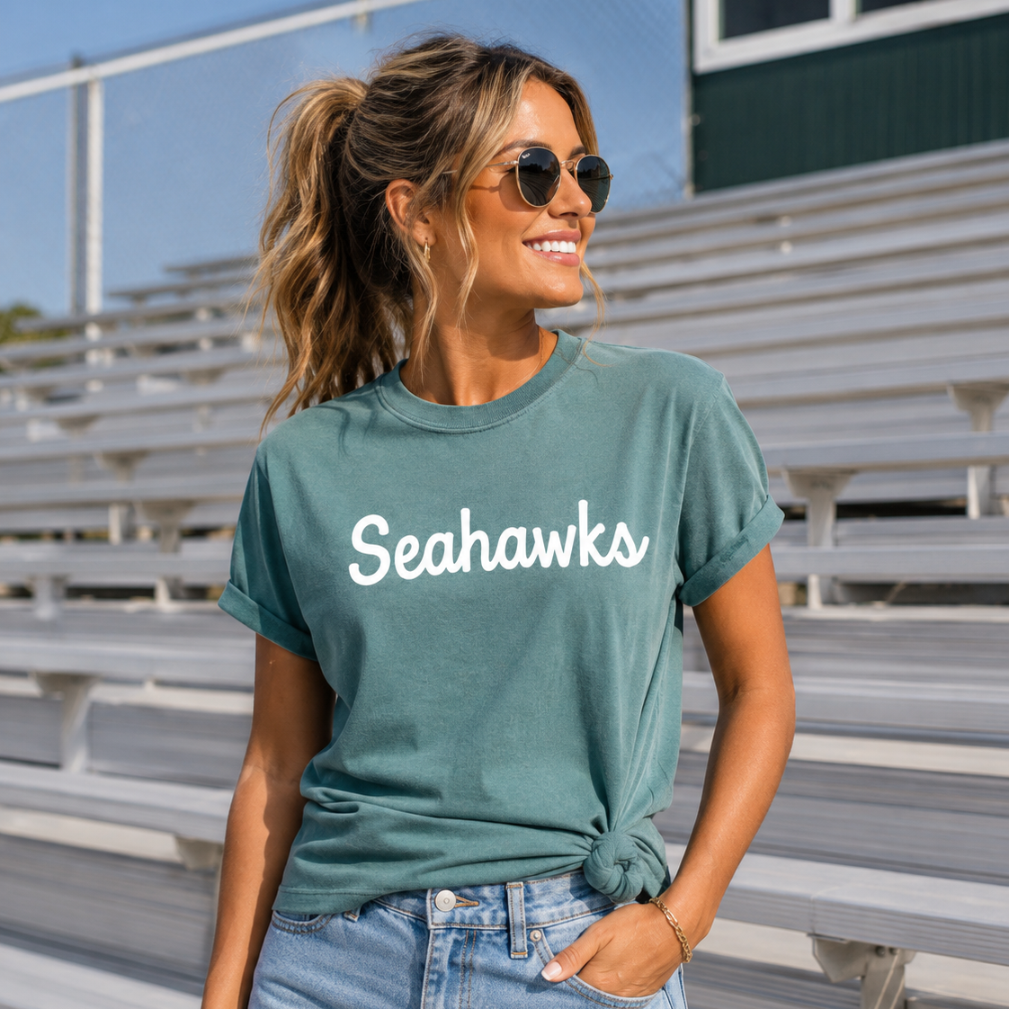A woman wearing a teal green short-sleeve t-shirt with 'Seahawks' printed in white script across the chest, paired with denim shorts, standing in front of outdoor stadium bleachers.