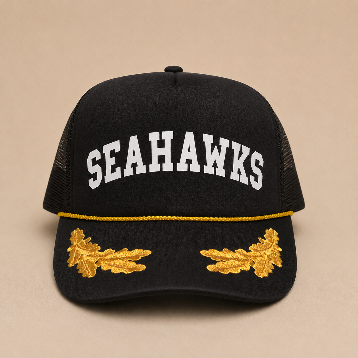 A black trucker hat with 'SEAHAWKS' embroidered in white arched letters on the front, a thin yellow rope detail across the brim, and gold embroidered oak leaves on each side of the brim, set against a solid beige background.