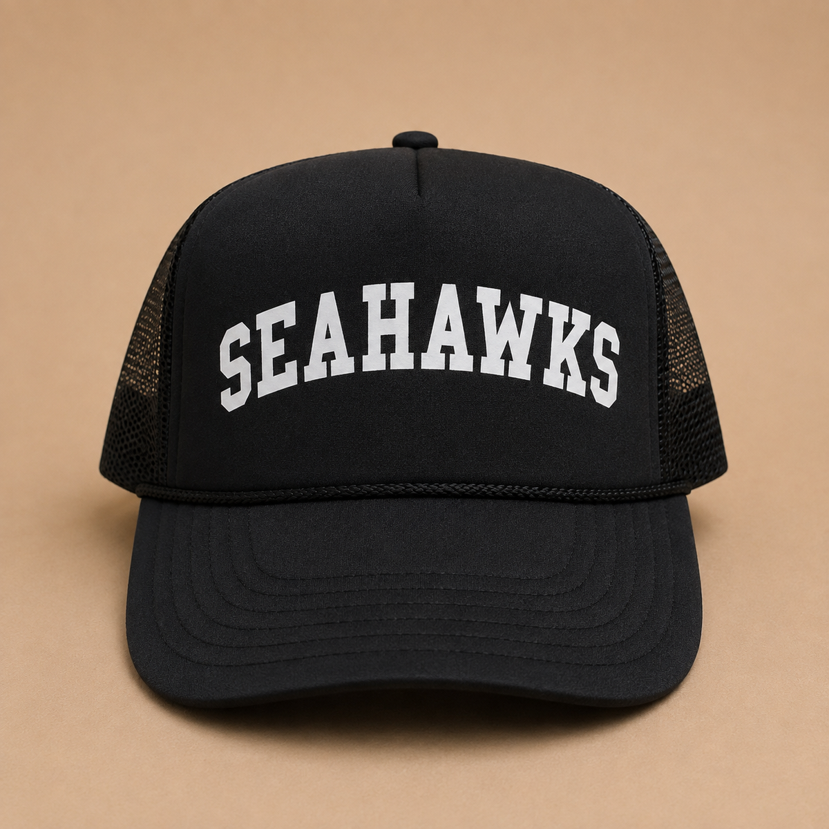 A black trucker-style baseball cap with 'SEAHAWKS' embroidered in white arched block letters on the front, set against a solid tan background.