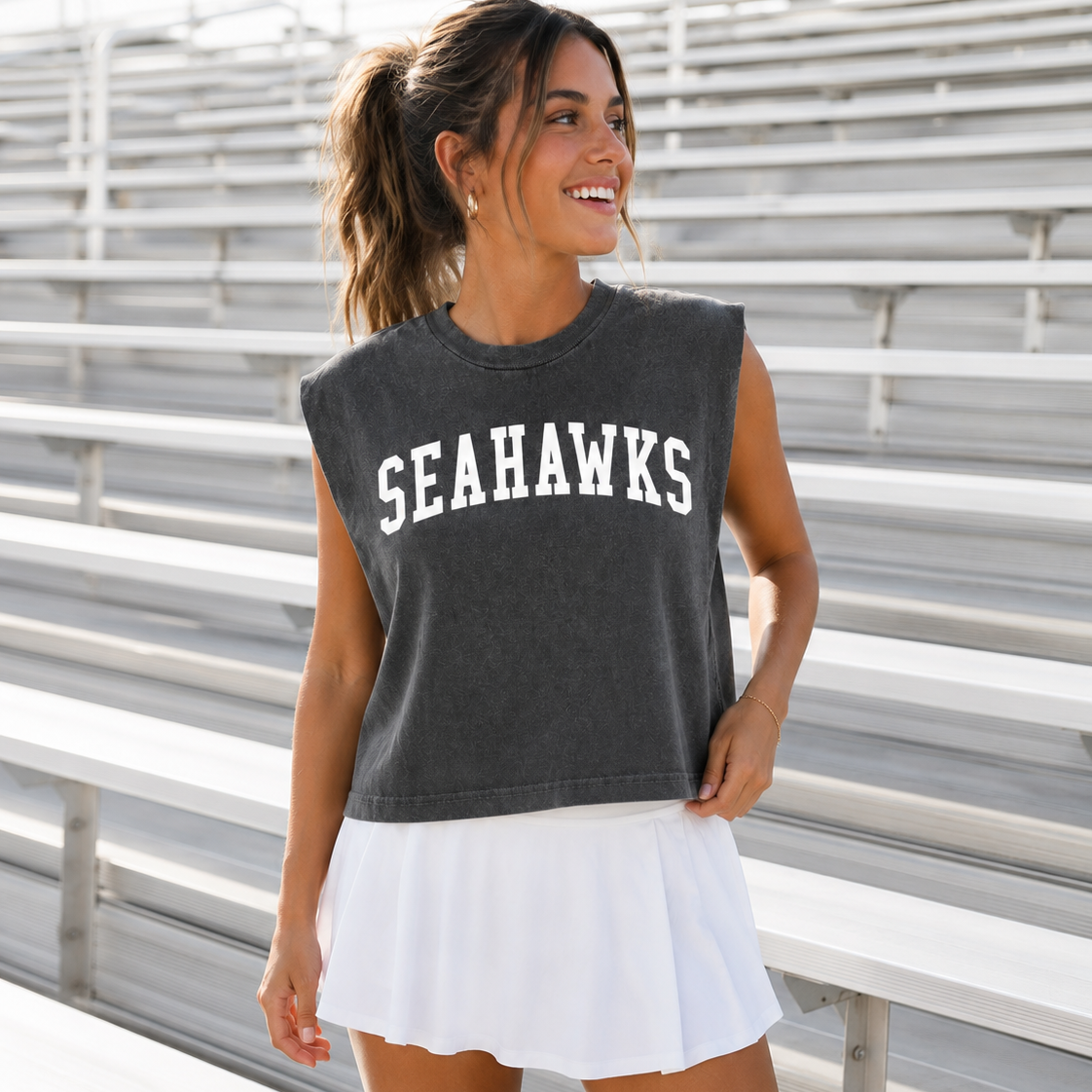 A woman wearing a dark gray sleeveless crop top with 'SEAHAWKS' printed in white across the chest, paired with a white tennis skirt, standing in front of outdoor stadium bleachers.