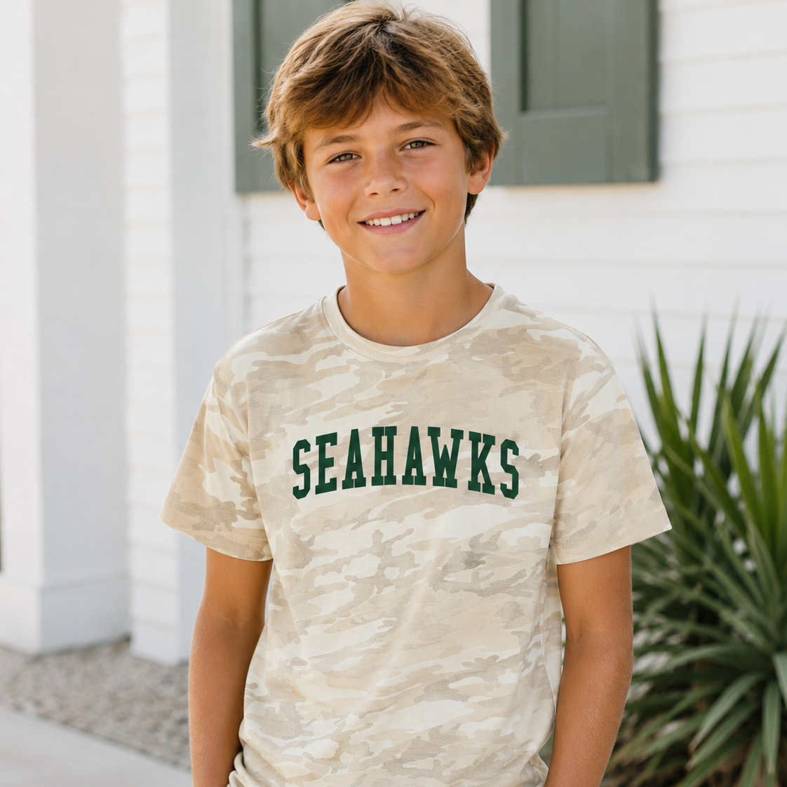 A smiling young boy wearing a beige camouflage t-shirt with 'SEAHAWKS' printed in dark green across the chest, paired with khaki shorts, standing outdoors in front of a white house.