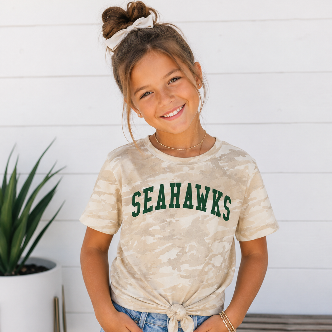 A young girl smiling and posing in a beige camouflage t-shirt with 'SEAHAWKS' printed in green across the chest, paired with light blue denim shorts, set against a white wood-paneled wall background.