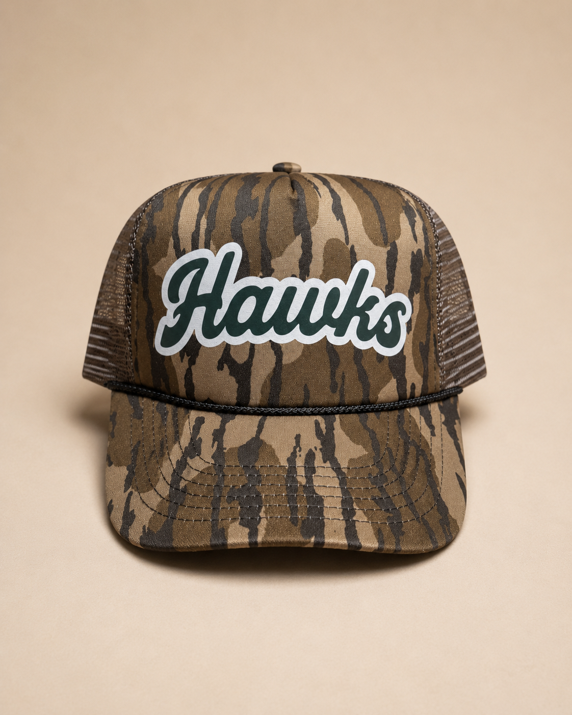 A camouflage trucker hat with a curved brim and mesh back, featuring the word 'Hawks' in white script with a black outline on the front panel, set against a solid beige background.