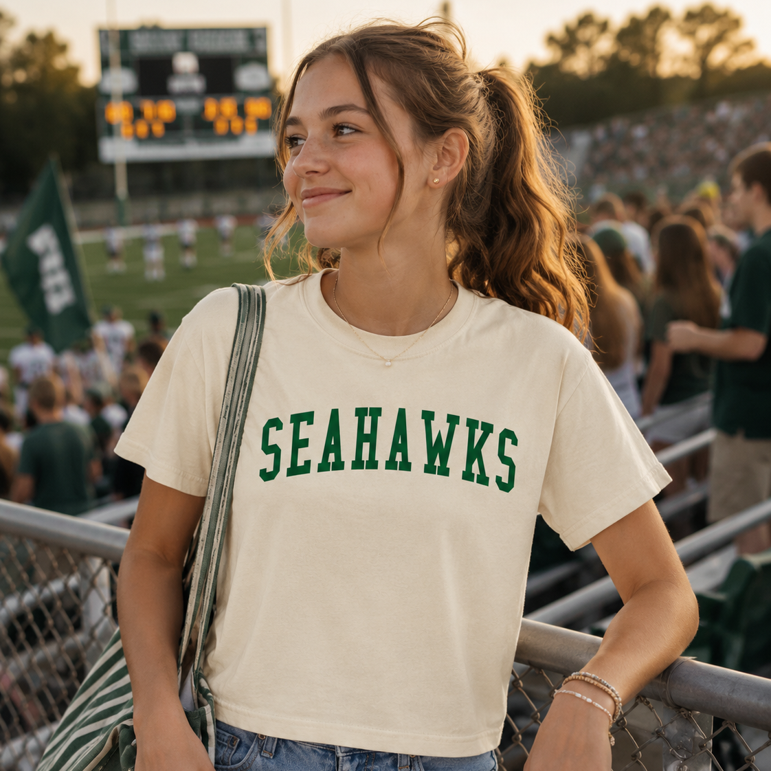 A woman at a stadium wearing a cream-colored short-sleeve t-shirt with 'SEAHAWKS' printed in green across the chest, paired with light blue jeans and a green striped shoulder bag.