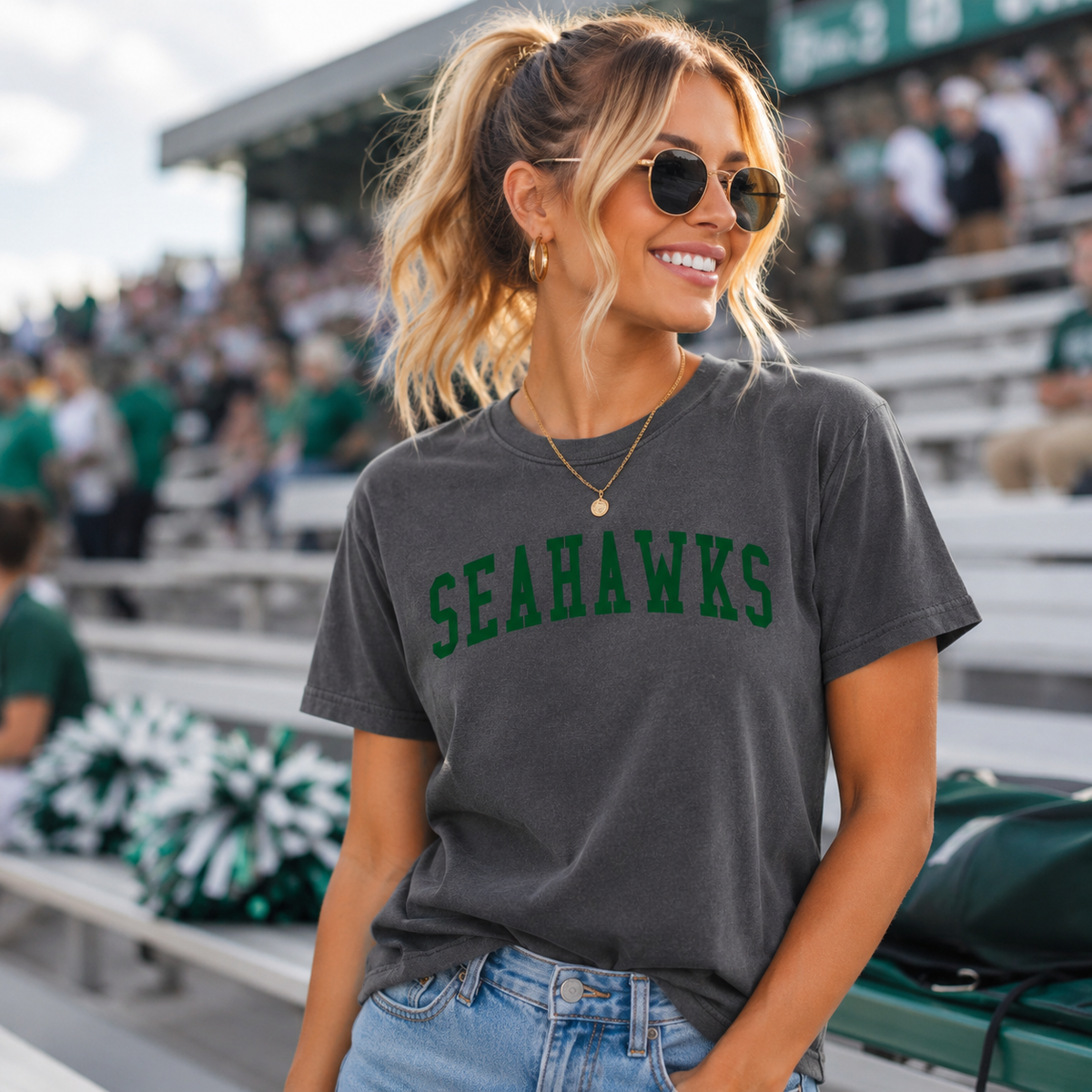 A woman wearing a dark gray Seahawks graphic t-shirt and blue jeans, standing in a stadium crowd.